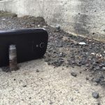 A bolt protruding from a road, close to the curb with a phone used to give scale